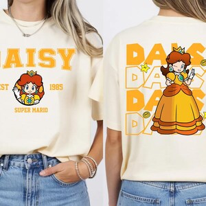 May include: Cream-colored t-shirt featuring the name "DAISY" in orange block letters, with a graphic of Princess Daisy from Super Mario. The back of the shirt repeats "DAISY" with a Princess Daisy graphic and stars.