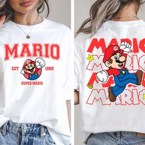 May include: White t-shirt featuring a front and back design of the video game character Mario. The front design includes the word "MARIO" in red block letters, the character's image, and the text "EST 1985 SUPER MARIO". The back design repeats the word "MARIO" and the character.