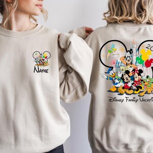 May include: Beige sweatshirt featuring Disney characters. The front has a Mickey Mouse head design with characters and the word "Name". The back displays a larger Mickey Mouse head design with characters, fireworks, the year "2020", and "Disney Family Vacation".