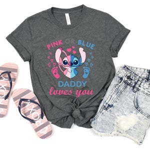 Puede incluir: Una camiseta gris con un gr&aacute;fico de Stitch rosa y azul. El texto "Pink or Blue Daddy loves you" est&aacute; impreso en la camiseta.