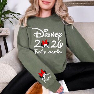 May include: Olive green sweatshirt with white text that reads "Disney 2020 Family Vacation". A Minnie Mouse graphic with a red bow is featured in the center and on the sleeve with the word "Name".
