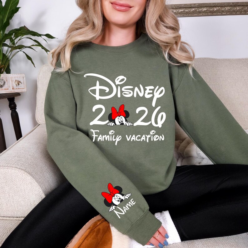 May include: Olive green sweatshirt with white text that reads "Disney 2020 Family Vacation". A Minnie Mouse graphic with a red bow is featured in the center and on the sleeve with the word "Name".