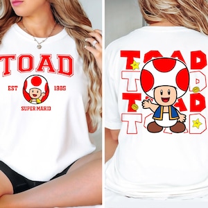 May include: White t-shirt featuring a red and white graphic of the character Toad from Super Mario. The front of the shirt has the word "TOAD" in red block letters, with "EST 1985" and "SUPER MARIO" below. The back has a larger Toad graphic and the word "TOAD" repeated.