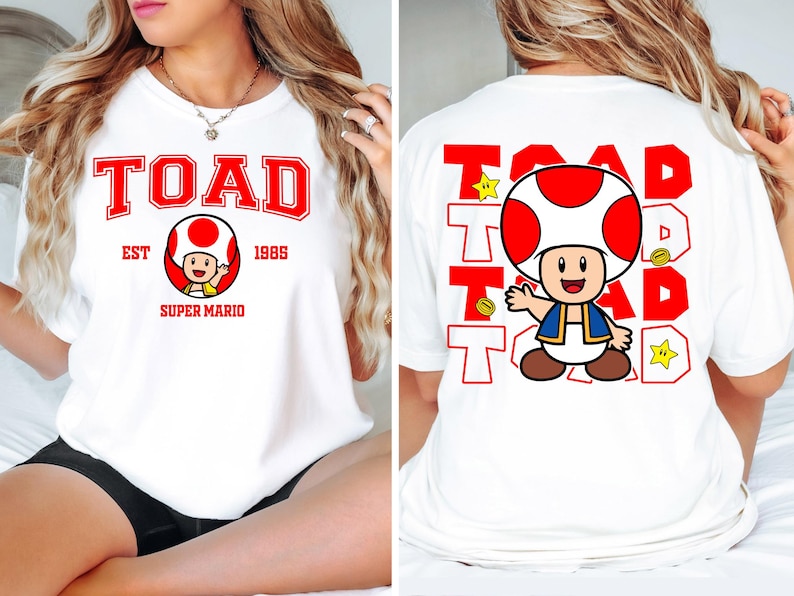 May include: White t-shirt featuring a red and white graphic of the character Toad from Super Mario. The front of the shirt has the word "TOAD" in red block letters, with "EST 1985" and "SUPER MARIO" below. The back has a larger Toad graphic and the word "TOAD" repeated.