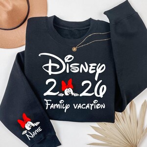 May include: Black sweatshirt with the text "Disney 2026 Family Vacation" in white script. Minnie Mouse with a red bow is featured in the design. The sleeve has a Minnie Mouse graphic and space for a name.
