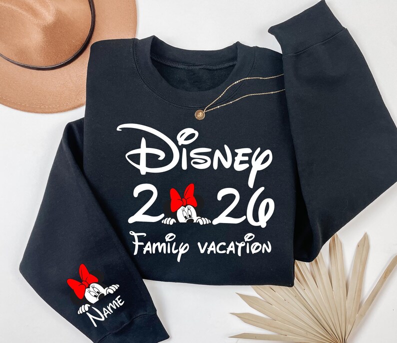 May include: Black sweatshirt with the text "Disney 2026 Family Vacation" in white script. Minnie Mouse with a red bow is featured in the design. The sleeve has a Minnie Mouse graphic and space for a name.