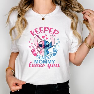 Puede incluir: Camiseta blanca con un personaje de Stitch azul y rosa sosteniendo una huella de pie de beb&eacute; rosa y azul. El texto en la camiseta dice "Keeper of the gender mommy loves you".
