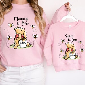 Peut inclure: Sweat-shirts roses assortis avec des illustrations de Winnie l'Ourson. L'un dit "Mommy to Bee", l'autre "Sister to Bee". Les deux pr&eacute;sentent Winnie avec un pot de miel et des abeilles.