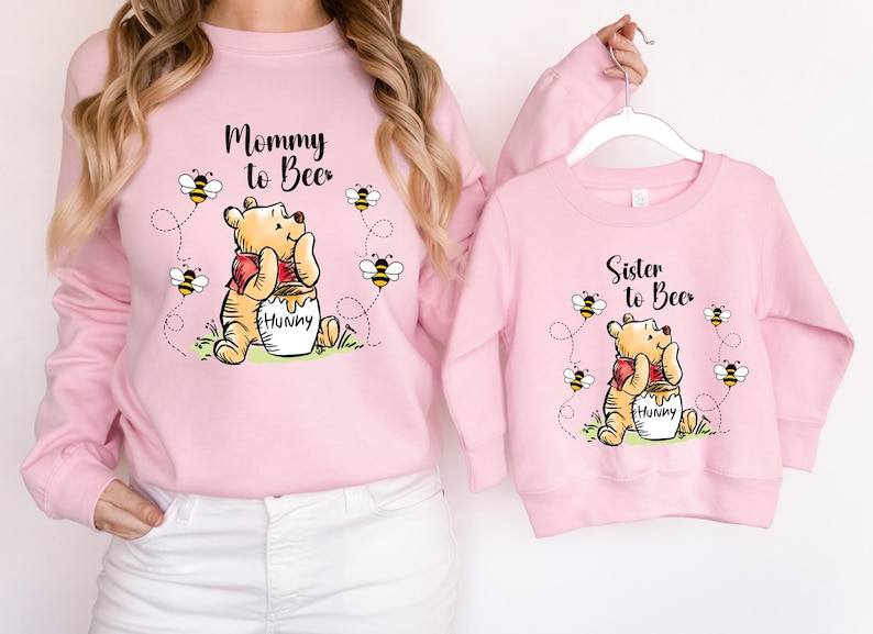 Peut inclure: Sweat-shirts roses assortis avec des illustrations de Winnie l'Ourson. L'un dit "Mommy to Bee", l'autre "Sister to Bee". Les deux pr&eacute;sentent Winnie avec un pot de miel et des abeilles.