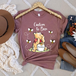 Peut inclure: T-shirt mauve avec un motif Winnie l'ourson, des abeilles et le texte "Custom to Bee". L'image comprend un chapeau marron, un jean et des chaussures plates marron, sur un tapis cr&egrave;me.