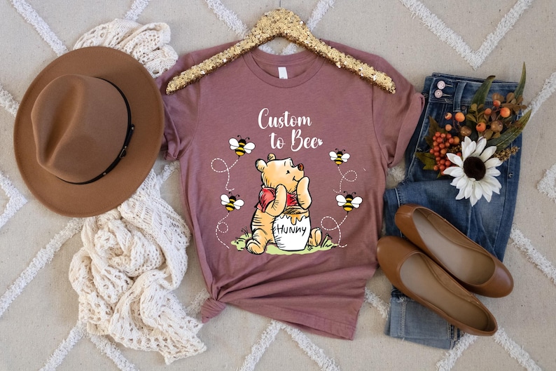Peut inclure: T-shirt mauve avec un motif Winnie l'ourson, des abeilles et le texte "Custom to Bee". L'image comprend un chapeau marron, un jean et des chaussures plates marron, sur un tapis cr&egrave;me.