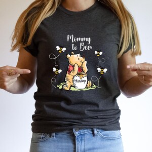 Peut inclure: T-shirt gris fonc&eacute; avec l'inscription "Mommy to Bee" et une illustration de Winnie l'Ourson avec un pot de miel et des abeilles. Le t-shirt est en mati&egrave;re souple, adapt&eacute; au port d&eacute;contract&eacute;.
