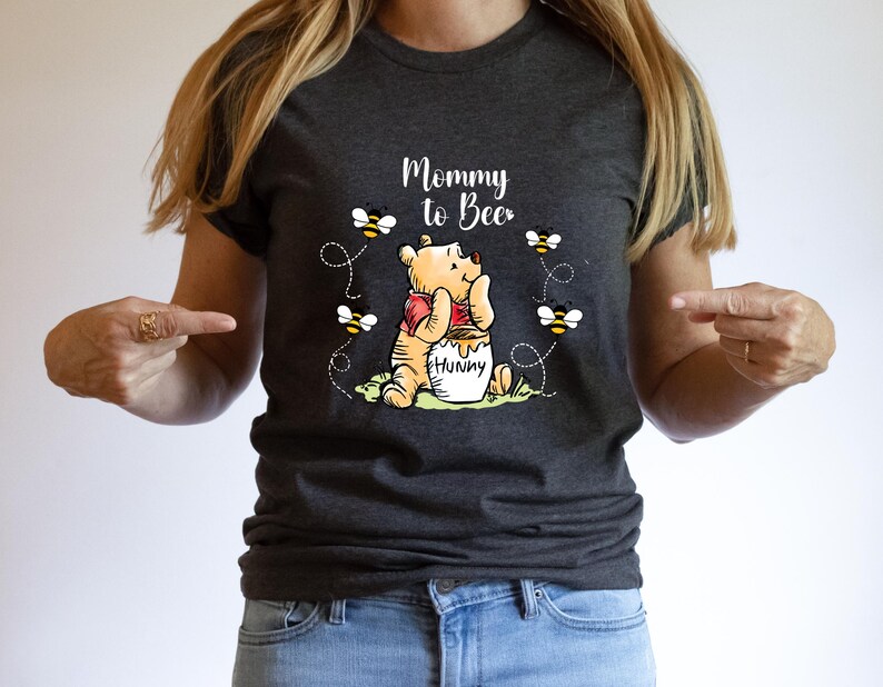 Peut inclure: T-shirt gris fonc&eacute; avec l'inscription "Mommy to Bee" et une illustration de Winnie l'Ourson avec un pot de miel et des abeilles. Le t-shirt est en mati&egrave;re souple, adapt&eacute; au port d&eacute;contract&eacute;.
