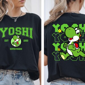 May include: Black t-shirt featuring a green Yoshi graphic from Super Mario. The front displays "YOSHI" in green block letters, with "EST 1985" and a Yoshi character illustration. The back shows a running Yoshi with "YOSHI" repeated.