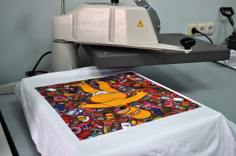 Peut inclure: Un t-shirt blanc avec un motif graphique color&eacute; repr&eacute;sentant un personnage de dessin anim&eacute; jaune entour&eacute; d'aliments. Le t-shirt est sur une presse &agrave; chaud, sugg&eacute;rant un processus d'impression personnalis&eacute;.