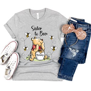 Peut inclure: T-shirt gris clair avec l'inscription "Sister to Bee" et un dessin de Winnie l'Ourson avec des abeilles. L'image comprend un jean bleu, un serre-t&ecirc;te &agrave; paillettes or rose et des baskets blanches.