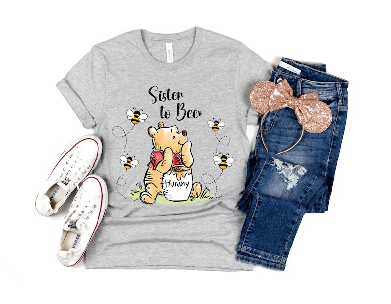 Peut inclure: T-shirt gris clair avec l'inscription "Sister to Bee" et un dessin de Winnie l'Ourson avec des abeilles. L'image comprend un jean bleu, un serre-t&ecirc;te &agrave; paillettes or rose et des baskets blanches.