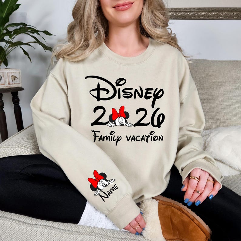 May include: Beige sweatshirt with black Disney logo, "2026", and "Family Vacation" text. Features a Minnie Mouse graphic with a red bow. The sleeve has a Minnie Mouse graphic and the word "Name".