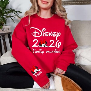 May include: Red sweatshirt with white text that reads "Disney 2020 Family Vacation." Minnie Mouse graphics are featured on the front and sleeve. The sleeve graphic includes a space for a name.