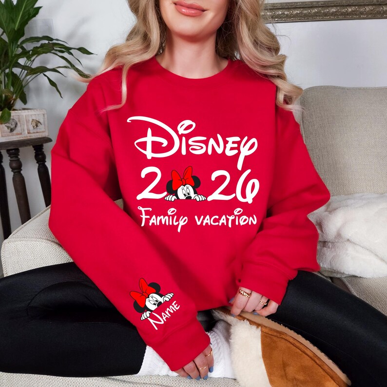 May include: Red sweatshirt with white text that reads "Disney 2020 Family Vacation." Minnie Mouse graphics are featured on the front and sleeve. The sleeve graphic includes a space for a name.