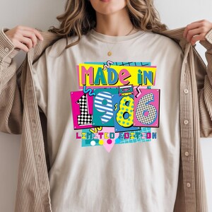 May include: Beige t-shirt with a colorful graphic that reads "MADE IN 1986 LIMITED EDITION." The design features a retro cassette tape, geometric shapes, and vibrant colors like pink, yellow, and blue. The shirt is being held open by a person.