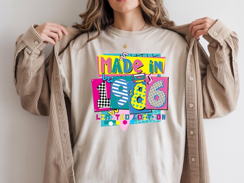 May include: Beige t-shirt with a colorful graphic that reads "MADE IN 1986 LIMITED EDITION." The design features a retro cassette tape, geometric shapes, and vibrant colors like pink, yellow, and blue. The shirt is being held open by a person.