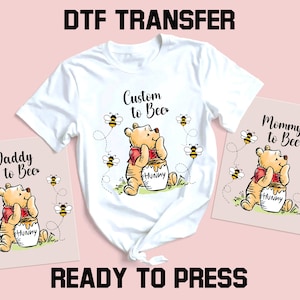 Peut inclure: T-shirt blanc avec un motif Winnie l'ourson, entour&eacute; d'abeilles, et le texte "Custom to Bee". Deux cartes roses avec le m&ecirc;me motif et le texte "Daddy to Bee" et "Mommy to Bee". Les mots "DTF TRANSFER" et "READY TO PRESS" sont &eacute;galement visibles.