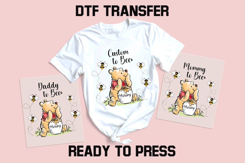 Peut inclure: T-shirt blanc avec un motif Winnie l'ourson, entour&eacute; d'abeilles, et le texte "Custom to Bee". Deux cartes roses avec le m&ecirc;me motif et le texte "Daddy to Bee" et "Mommy to Bee". Les mots "DTF TRANSFER" et "READY TO PRESS" sont &eacute;galement visibles.