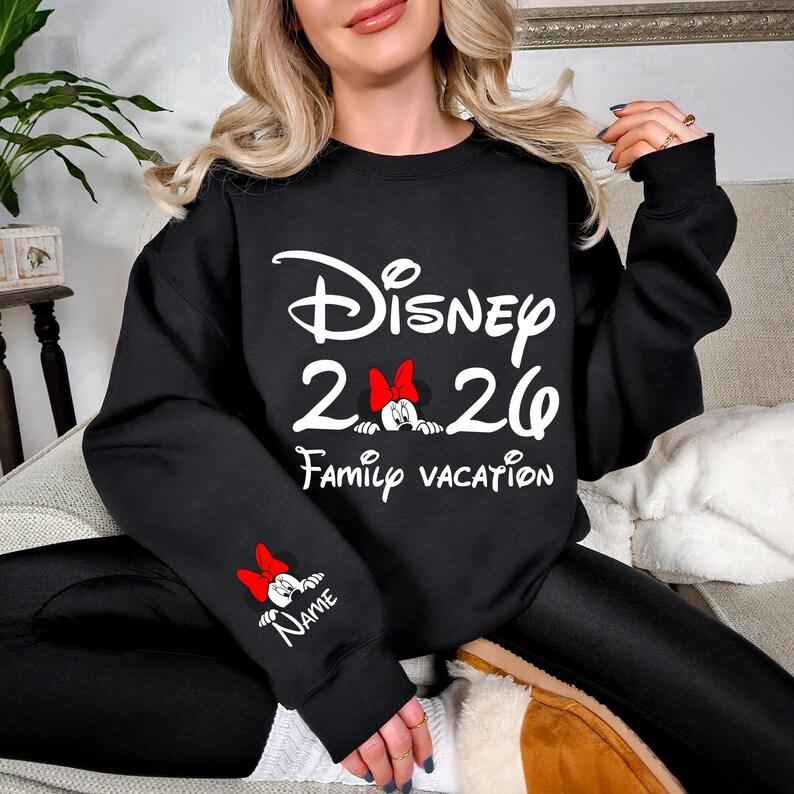 May include: Black sweatshirt with white text that reads "Disney 2026 Family Vacation." The design includes a Minnie Mouse graphic with a red bow. A smaller Minnie Mouse graphic is on the sleeve with the word "Name" below.