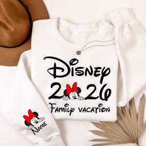 May include: White crewneck sweatshirt with black text that reads "Disney 2026 Family Vacation". Minnie Mouse graphic with a red bow is featured. A smaller Minnie Mouse graphic is on the sleeve with the word "Name".
