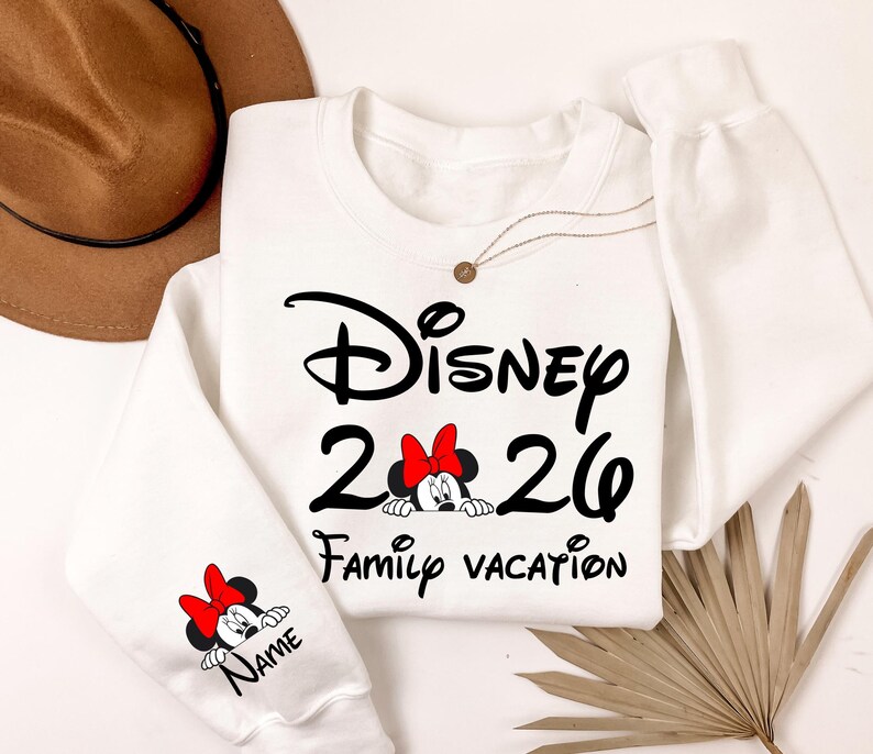 May include: White crewneck sweatshirt with black text that reads "Disney 2026 Family Vacation". Minnie Mouse graphic with a red bow is featured. A smaller Minnie Mouse graphic is on the sleeve with the word "Name".