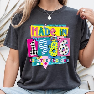 May include: Dark gray t-shirt with a colorful graphic that reads "MADE IN 1986 LIMITED EDITION." The design features the numbers 1, 9, 8, and 6 in vibrant colors and patterns, with a cassette tape illustration.