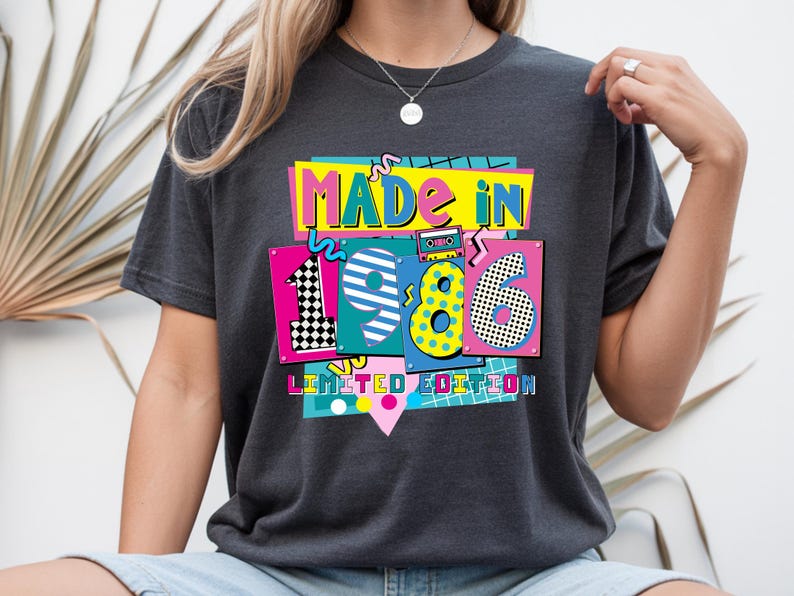 May include: Dark gray t-shirt with a colorful graphic that reads "MADE IN 1986 LIMITED EDITION." The design features the numbers 1, 9, 8, and 6 in vibrant colors and patterns, with a cassette tape illustration.