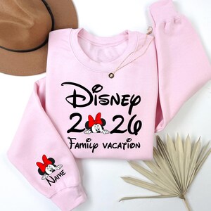 May include: A light pink sweatshirt with the word "Disney" in black script, the numbers "26", and the words "Family Vacation". A Minnie Mouse graphic with a red bow is featured. The sleeve has a Minnie Mouse graphic and the word "Name".