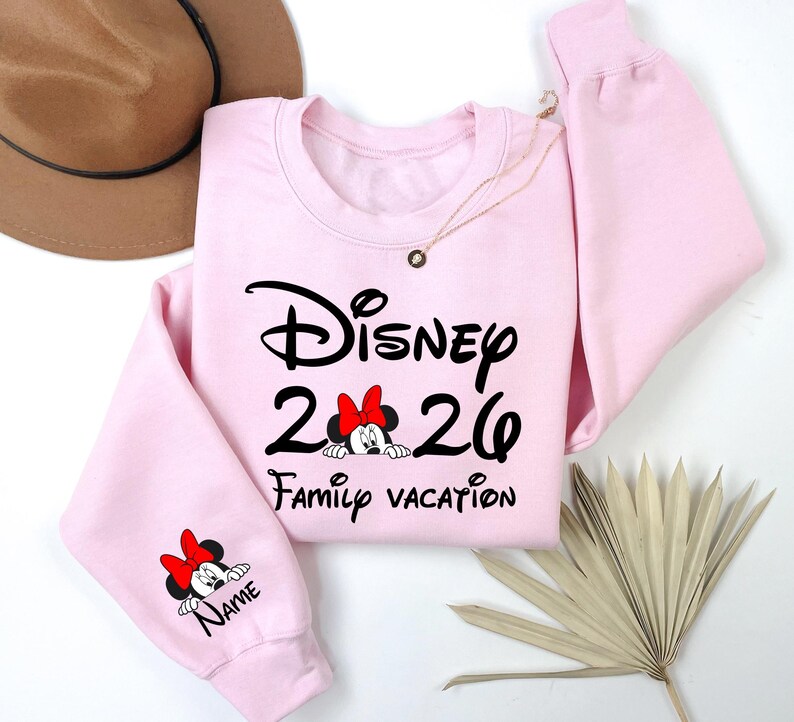 May include: A light pink sweatshirt with the word "Disney" in black script, the numbers "26", and the words "Family Vacation". A Minnie Mouse graphic with a red bow is featured. The sleeve has a Minnie Mouse graphic and the word "Name".
