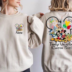 May include: Beige sweatshirt featuring a Mickey Mouse ear design with Disney characters. The front has a small design with the name, and the back has a larger design with the text "Family Vacation (Custom Text)" and the year 2026.