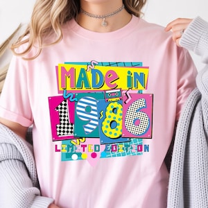 May include: Pink t-shirt with a colorful graphic design. The design features the words "MADE IN 1986 LIMITED EDITION" in a retro style, with vibrant colors and patterns. A cassette tape graphic is also included.