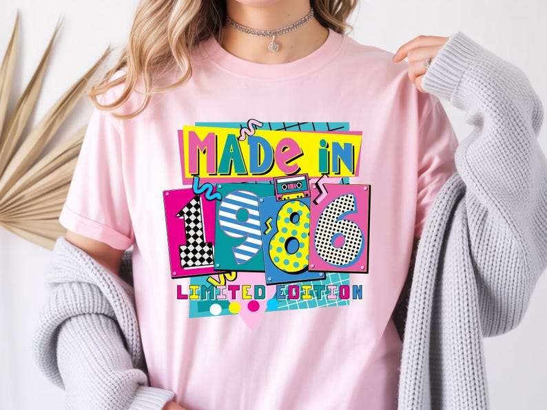 May include: Pink t-shirt with a colorful graphic design. The design features the words "MADE IN 1986 LIMITED EDITION" in a retro style, with vibrant colors and patterns. A cassette tape graphic is also included.