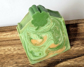 May include: A handmade soap bar with a green and white swirl pattern and a shamrock design on top. The soap has a light orange accent color and is displayed on a wooden surface.