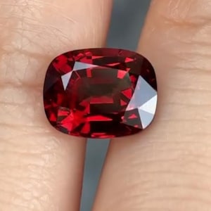 May include: A faceted, cushion-cut gemstone with a deep red hue. The gem displays multiple facets that reflect light, creating a brilliant sparkle. The shape is a rounded square, and the color is a rich, saturated red.