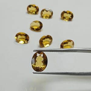 Può includere: Diverse gemme di citrino giallo dorato di forma ovale. Una gemma è tenuta da una pinzetta, mentre altre sono sparse su una superficie bianca. Le gemme hanno un taglio sfaccettato. Adatte per la creazione di gioielli.