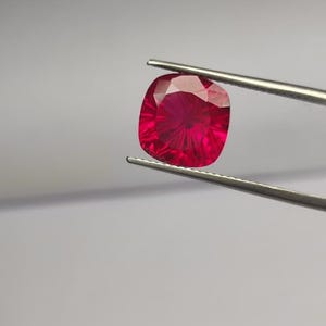 May include: A radiant, cushion-cut ruby gemstone, held by silver tweezers against a neutral backdrop. The gem's deep red hue and internal facets create a captivating display of light. Suitable for jewellery making.