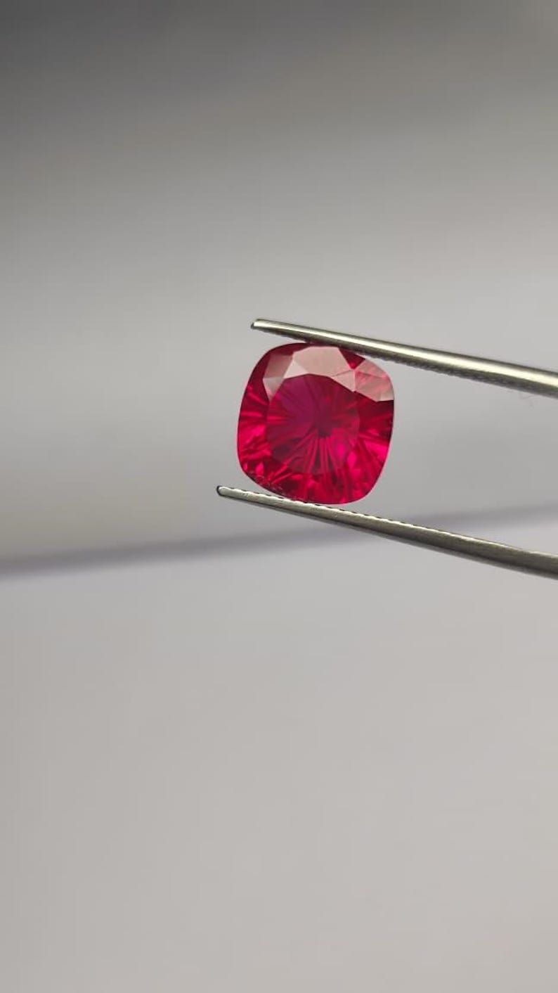 May include: A radiant, cushion-cut ruby gemstone, held by silver tweezers against a neutral backdrop. The gem's deep red hue and internal facets create a captivating display of light. Suitable for jewellery making.