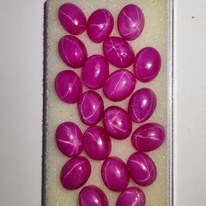 Op de afbeelding: Een verzameling ovale, magenta edelstenen met een ster-effect, gepresenteerd in een transparante plastic doos. De edelstenen zijn gerangschikt op een lichtgele schuimbasis. Het ster-effect wordt gecreëerd door elkaar kruisende lijnen.