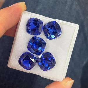 Op de afbeelding: Vijf briljantblauwe edelstenen met een kussensnede worden gepresenteerd in een wit bakje. De edelstenen hebben een rijke, verzadigde kleur en een gefacetteerd oppervlak dat licht reflecteert. Waarschijnlijk saffieren of vergelijkbare edelstenen.