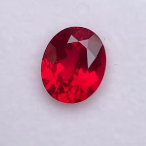 May include: A faceted, oval-shaped, deep red gemstone. The gem has a brilliant, reflective surface, showcasing multiple facets that catch and reflect light. The stone is set against a white background.