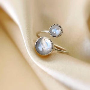 Op de afbeelding: Een zilveren ring met twee maansteen cabochons. Eén maansteen is groter en rond, de andere kleiner met een geschulpte bezel. De ring ligt op een crèmekleurige stof.