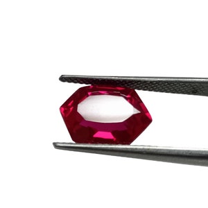 May include: A deep red gemstone with a faceted octagonal shape, held by tweezers against a white background.