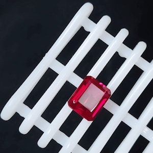 May include: A vibrant, rectangular-cut red gemstone with faceted surfaces, positioned centrally on a white plastic grid. The stone displays a deep, saturated crimson hue and clear transparency, reflecting light across its polished, geometric edges.