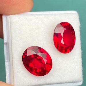 May include: Two faceted, oval-shaped, bright red gemstones are displayed in a white, rectangular container. The gems have a high luster and are set against a white background. The gemstones are a vibrant, saturated red color.